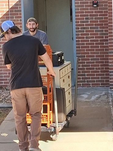 Two men moving furniture on a dolly outside a brick building.