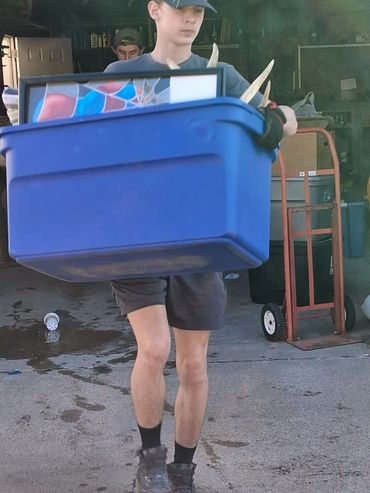 Young person carrying a blue bin with a framed artwork and antlers inside.