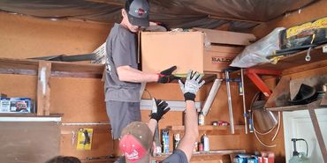 Two men passing a box in a cluttered workshop while a woman watches.
