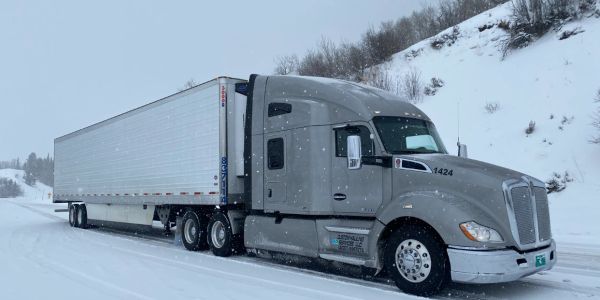 A gray semi-truck parked on a snowy road with snow-covered hills.