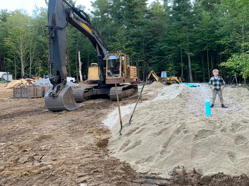 Young boy standing on top of a sand bed for a new septic system next to a 30 ton excavator