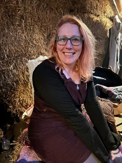 Smiling woman with glasses sitting against a hay bale in warm lighting.