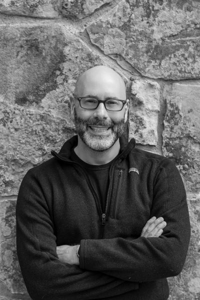 A man with glasses and a beard smiling, wearing a black sweater against a stone background.