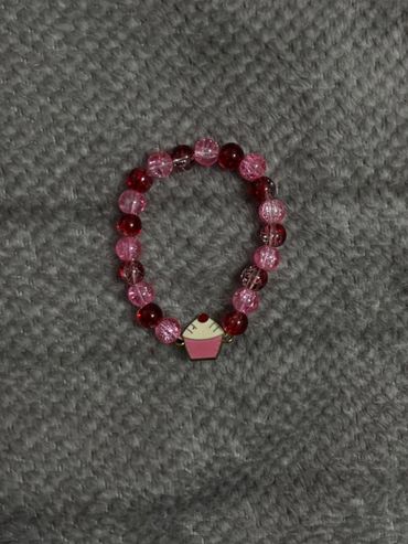 A pink and red beaded bracelet with a cupcake charm on a gray textured surface.