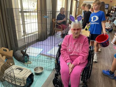 An elderly woman wearing bunny ears on her head sitting next to a rabbit pen