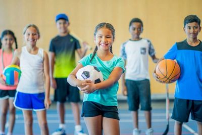 Smiling kids holding different sports balls in a gym.