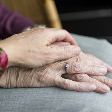 Person's hand on top of elderly person's hands, folded on lap.