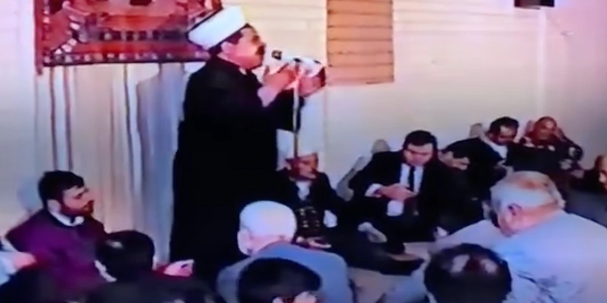 Man in traditional attire speaking to a seated audience indoors.