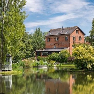 Brick house by a serene pond surrounded by lush greenery.