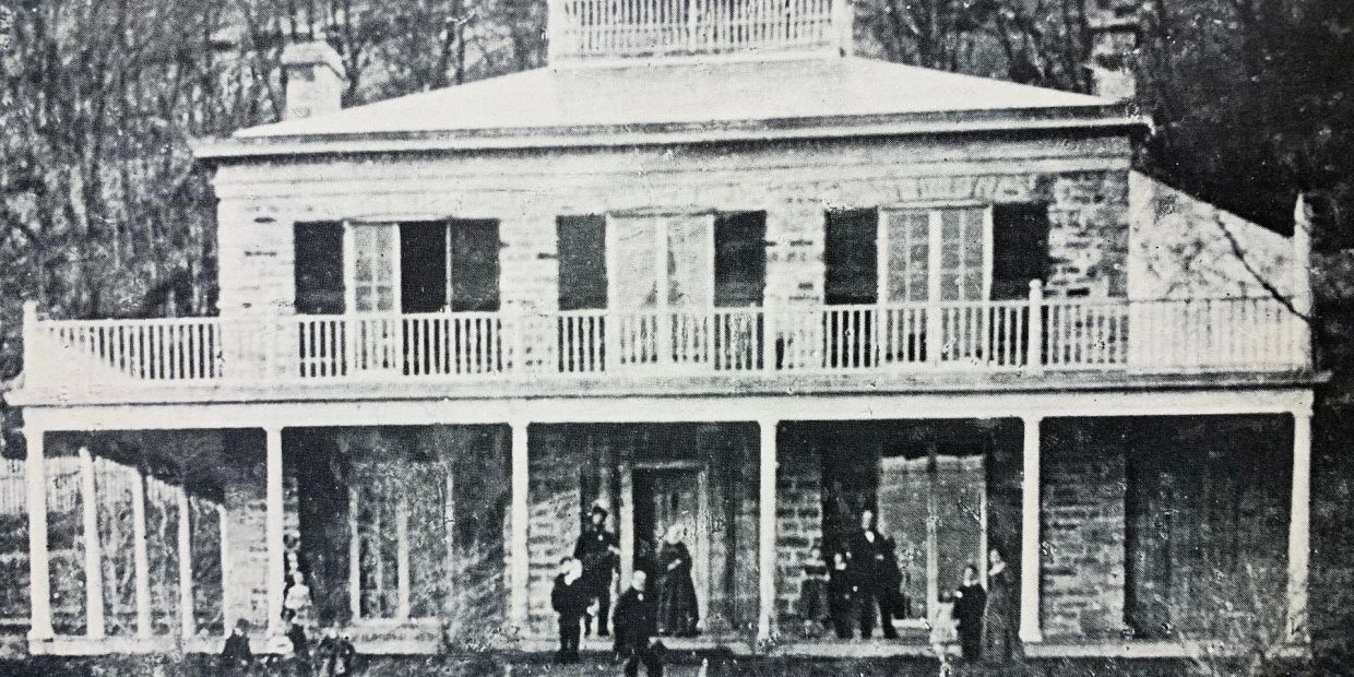 Old black and white photo of a large house with people in front.