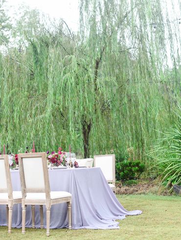 Elegant outdoor dining setup with lavender tablecloth and floral centerpiece under a weeping willow tree.