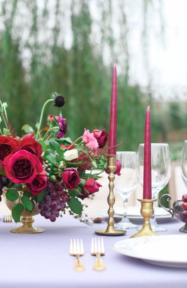 Elegant table setting with red roses, grapes, and tall red candles in brass holders.