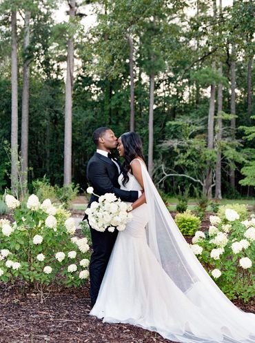 Bride and groom share a tender moment surrounded by white flowers and trees.