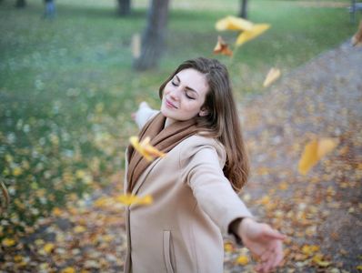 A woman with her eyes closed stands in a park with arms outstretched, surrounded by falling leaves.