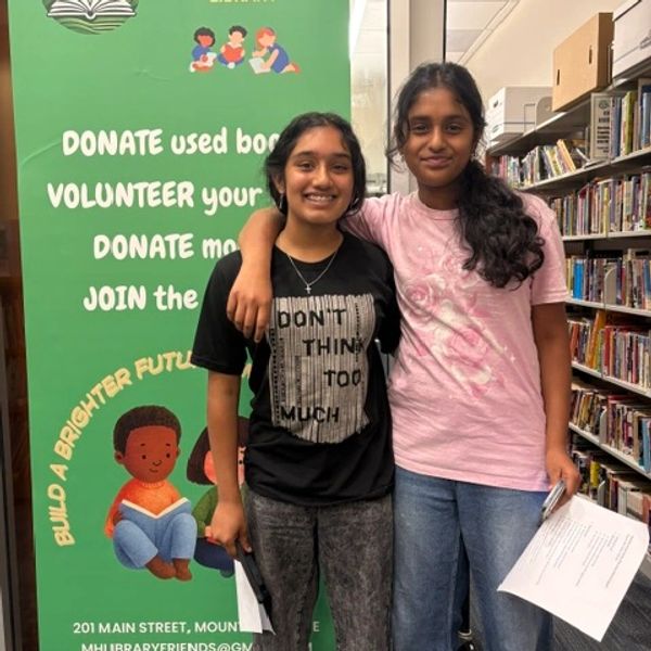 Two young women smiling in a library with a donation sign behind.