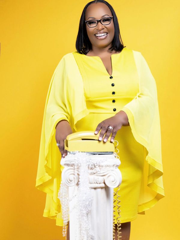Smiling woman in yellow dress with vintage phone on pillar.