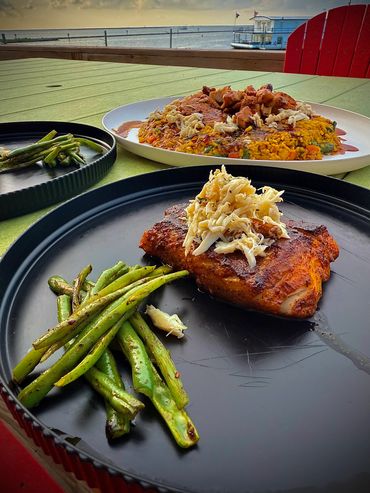 Grilled fish with crab meat and green beans on a black plate by the waterfront.