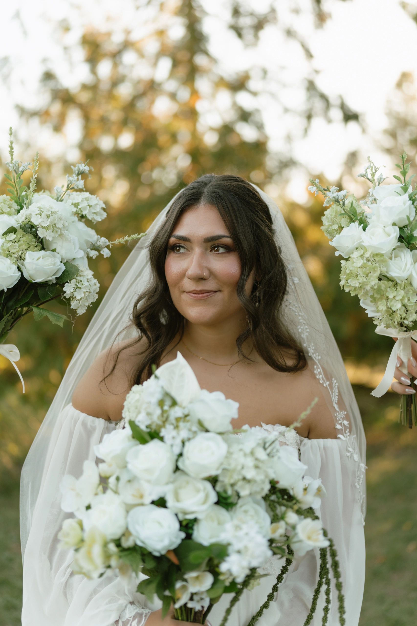 Bride holding white floral bouquets with a serene smile outdoors.