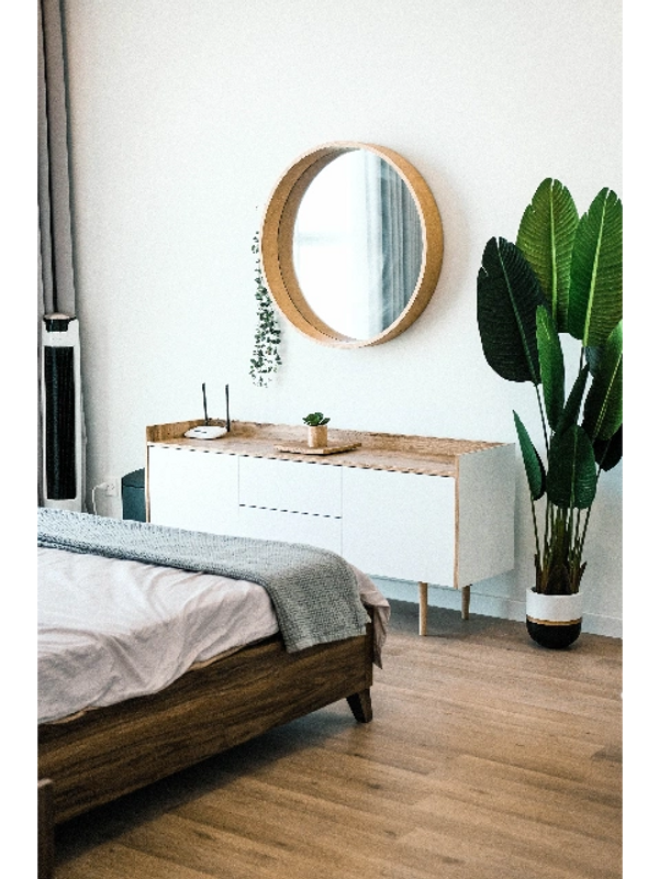 Minimalist bedroom corner with wooden bed, white cabinet, round mirror, and large green plant.