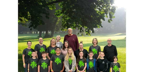 Group photo of children and adults in a sunny park with many wearing matching green logo shirts.