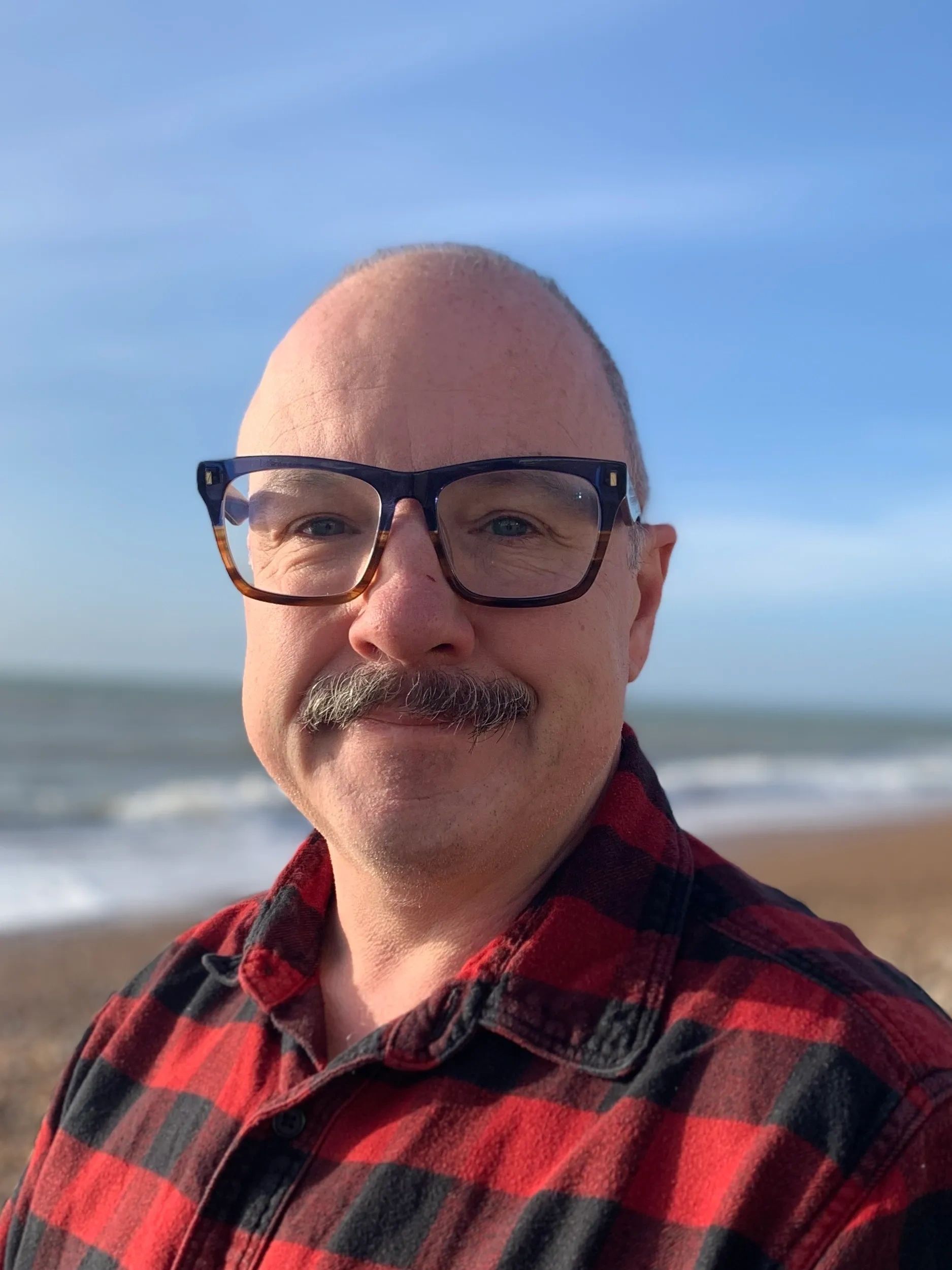 Smiling man with glasses and mustache in red plaid shirt at the beach.