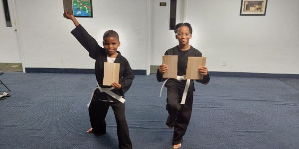 Two children in martial arts uniforms holding wooden boards, posing confidently indoors.