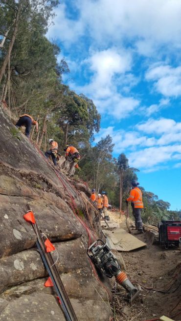 Workers in orange safety gear on rocky terrain with equipment and trees.
