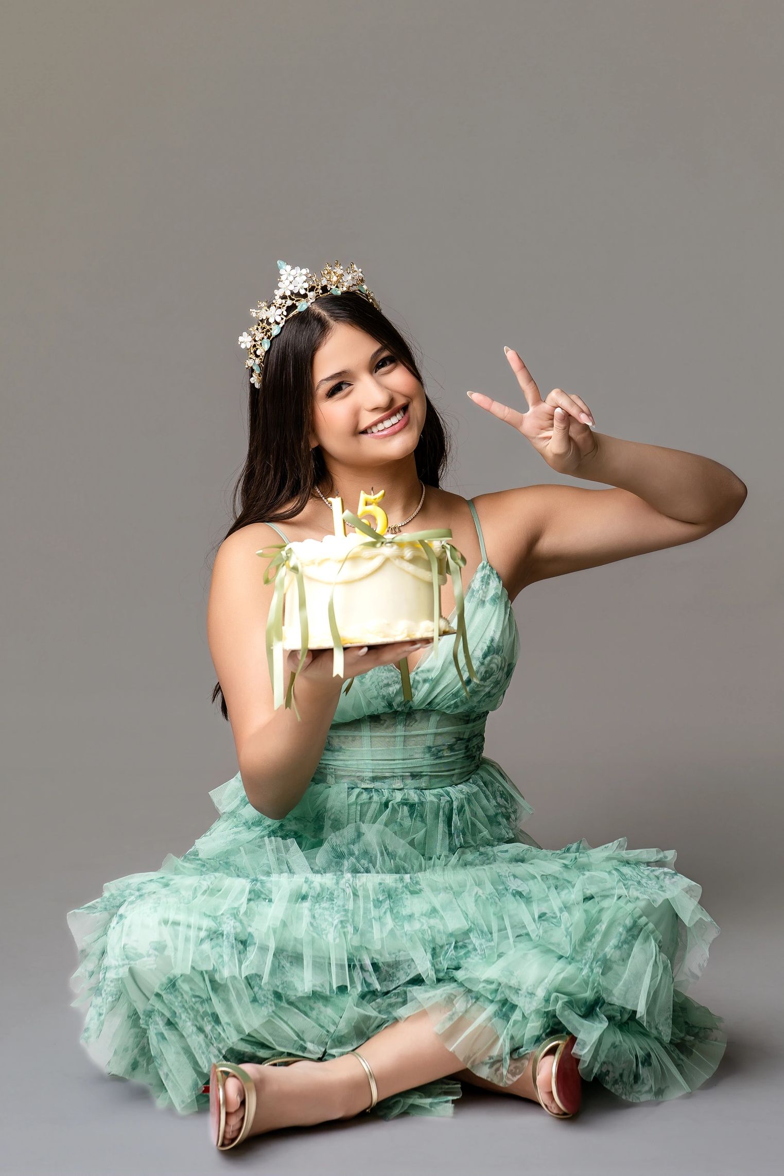 Young woman in green dress holding a 15th birthday cake and flashing a peace sign.