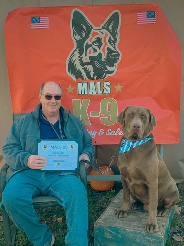Man holding a certification with his dog in front of a MALS K9 banner.