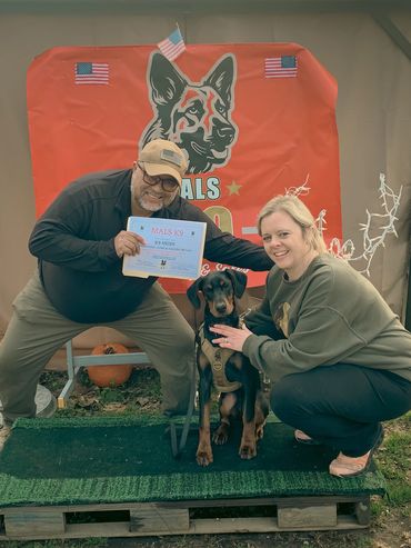 Two people proudly pose with a Doberman and a K9 training certificate.