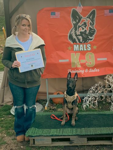 Woman holding a K9 certification with a trained dog in front of a MALS K-9 banner.