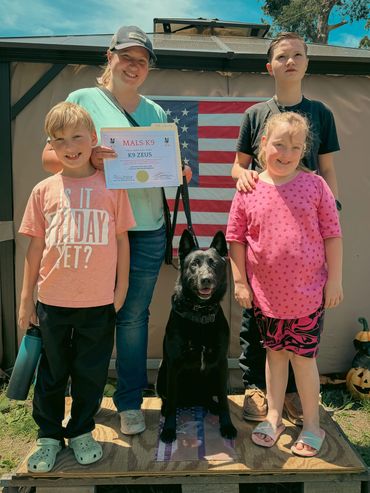 Family proudly posing with a certificate and their black dog in front of an American flag.