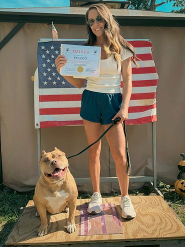 Woman proudly shows K9 Coco's training certificate with dog in front of American flag.