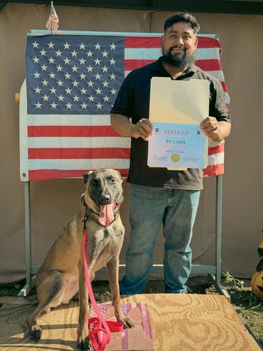 Man proudly holds a certificate for K9 Cora with his dog sitting beside him.