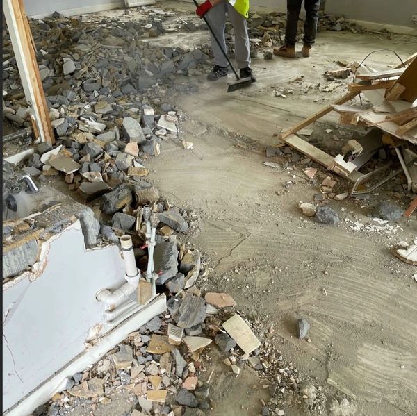 Workers cleaning debris from a demolished room with exposed pipes and rubble.