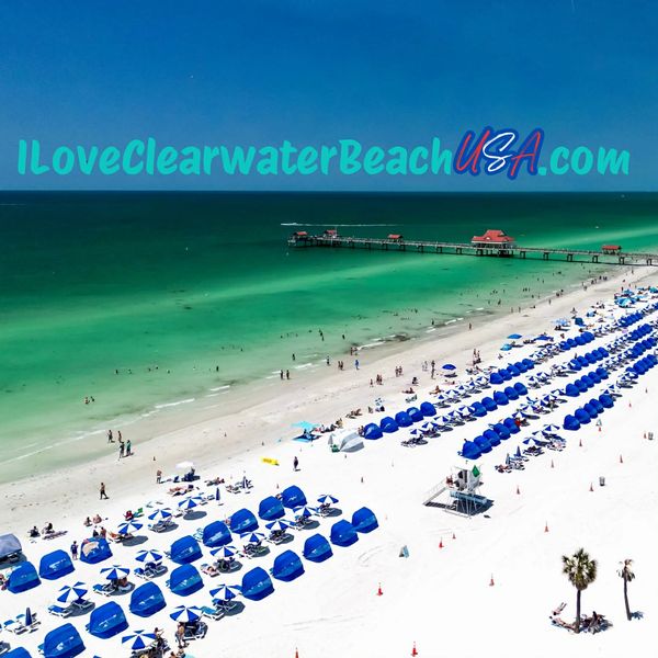 Aerial view of Clearwater Beach with blue umbrellas and a pier extending into clear green water.