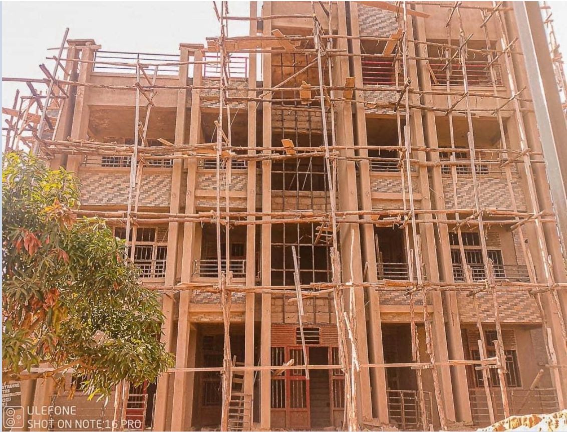 Multi-story building under construction with wooden scaffolding and surrounding trees.