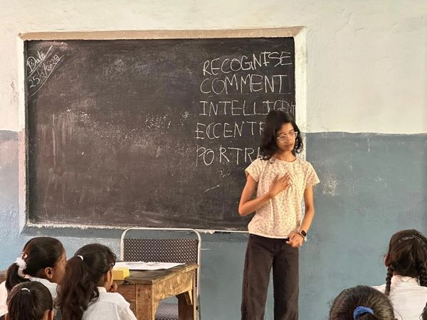 A teacher stands in front of a chalkboard with students seated, engaging in a lesson.