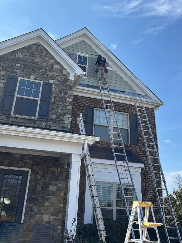 A worker on a ladder repairing or inspecting a house exterior under a clear sky.