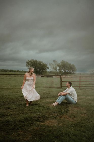 A woman in a floral dress dances while a man sits barefoot on grass under a cloudy sky.