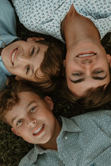 Three smiling boys lying head-to-head on grass, wearing patterned shirts.
