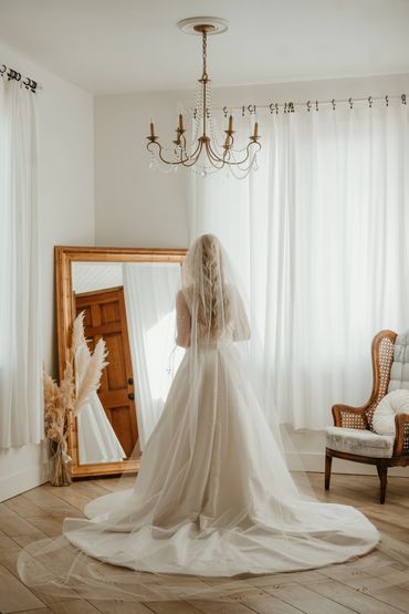 Bride in a flowing white gown gazes into a large mirror in a softly lit room.
