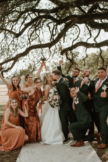 Bride and groom kiss surrounded by cheerful bridesmaids and groomsmen under a large tree.