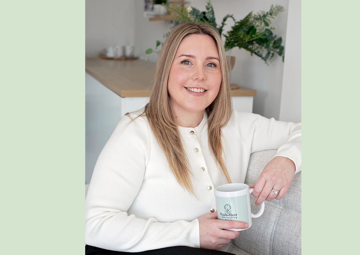 Smiling woman in white sweater holding a branded mug, sitting on a couch.