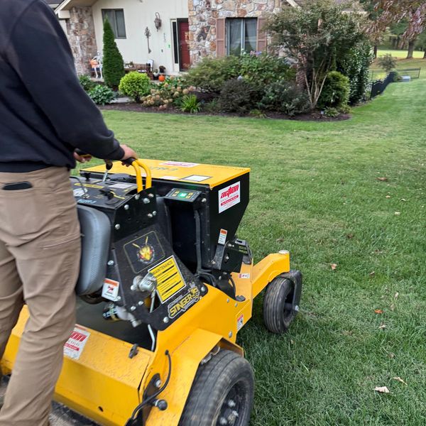 Person operating yellow lawn equipment near a house with a stone facade.