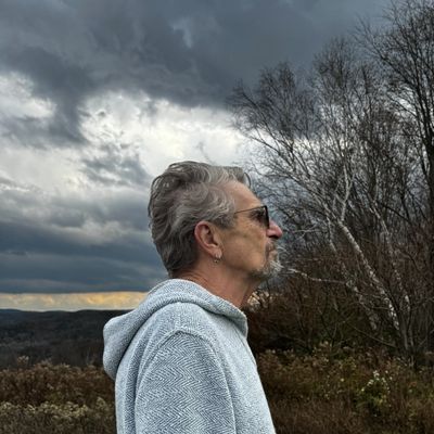Man with gray hair and glasses standing outdoors under a cloudy sky.