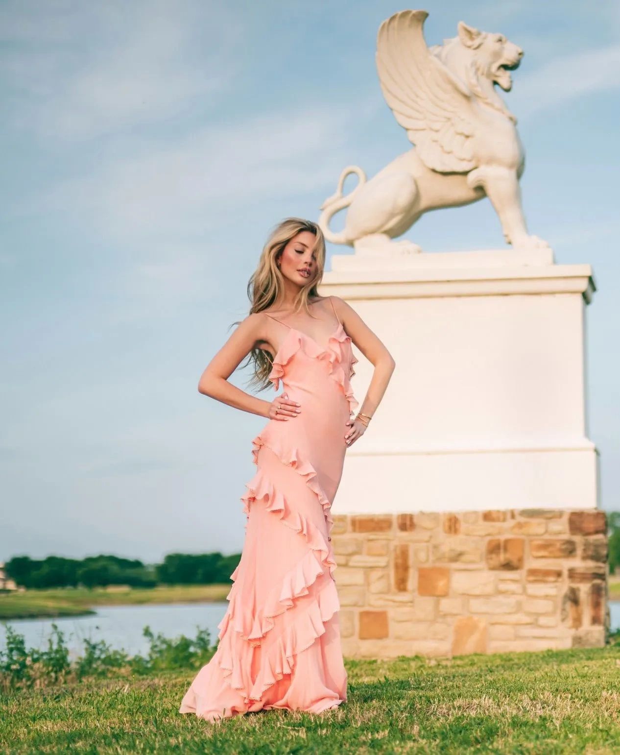 Woman in a flowing peach dress poses by a winged lion statue outdoors.