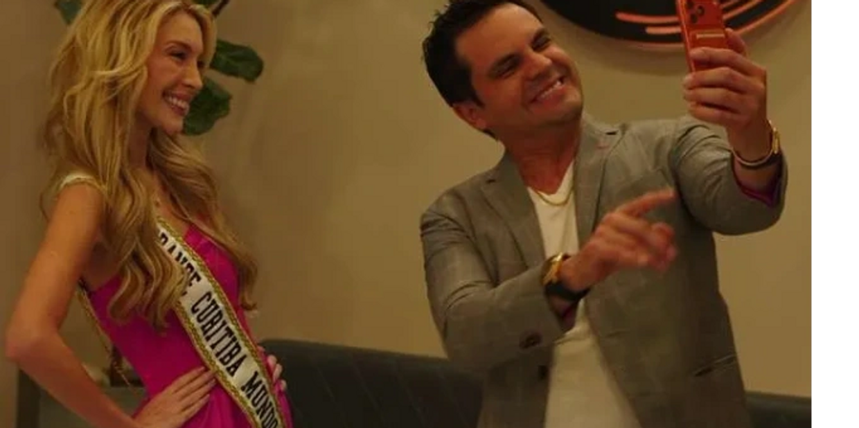 A man takes a selfie with a woman wearing a pink dress and a sash.