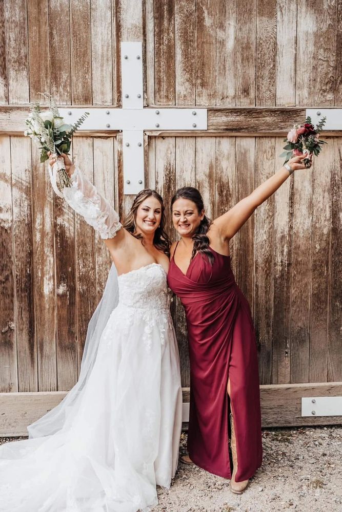 Bride and bridesmaid joyfully pose with bouquets in front of rustic wooden doors.