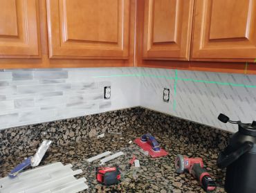 Kitchen backsplash installation with tools on countertop and wooden cabinets above.